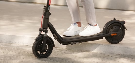 Osoba w białych butach stoi na czarnej hulajnodze NAVEE V25i Pro II, a obok widać krawężnik.